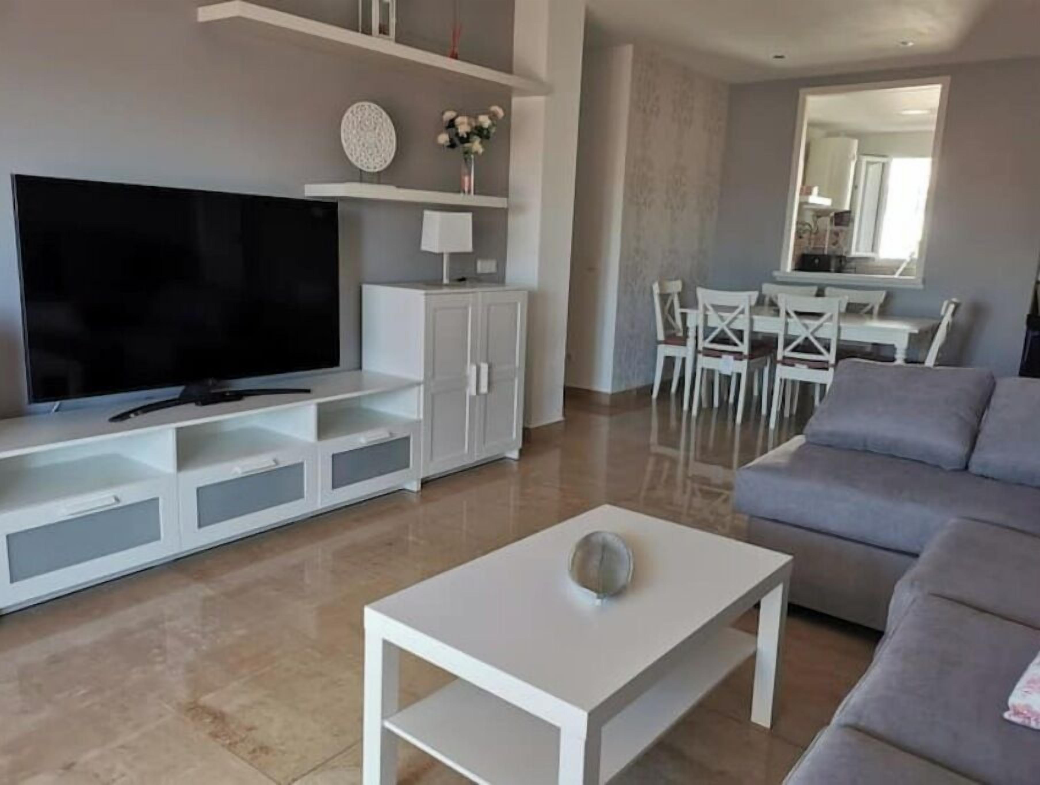 A modern living room with a flat-screen TV, white cabinets, a grey sofa, and a dining area with white chairs.