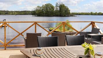 Una terraza de madera con muebles de mimbre y una mesa, con vista a un lago que tiene un muelle y árboles al fondo.