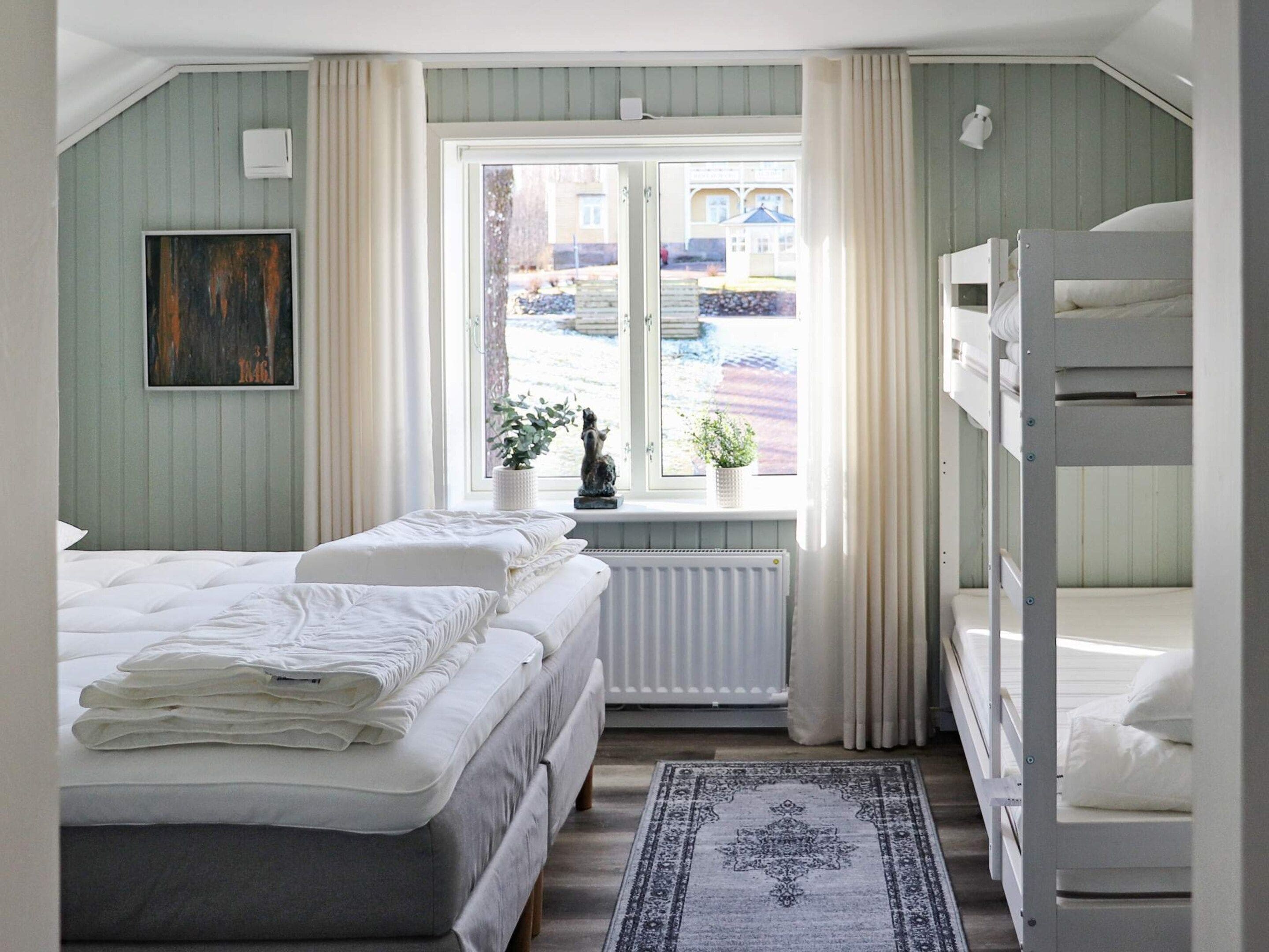 A bedroom with a bunk bed, a single bed, a window with curtains, a radiator, and a rug.