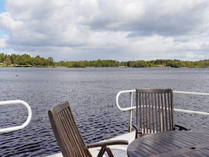 A boat deck with a wooden chair and table, a railing, and a view of a calm lake with trees and buildings in the distance. - 5 Star Holiday Home in Unnaryd (Unnaryd)