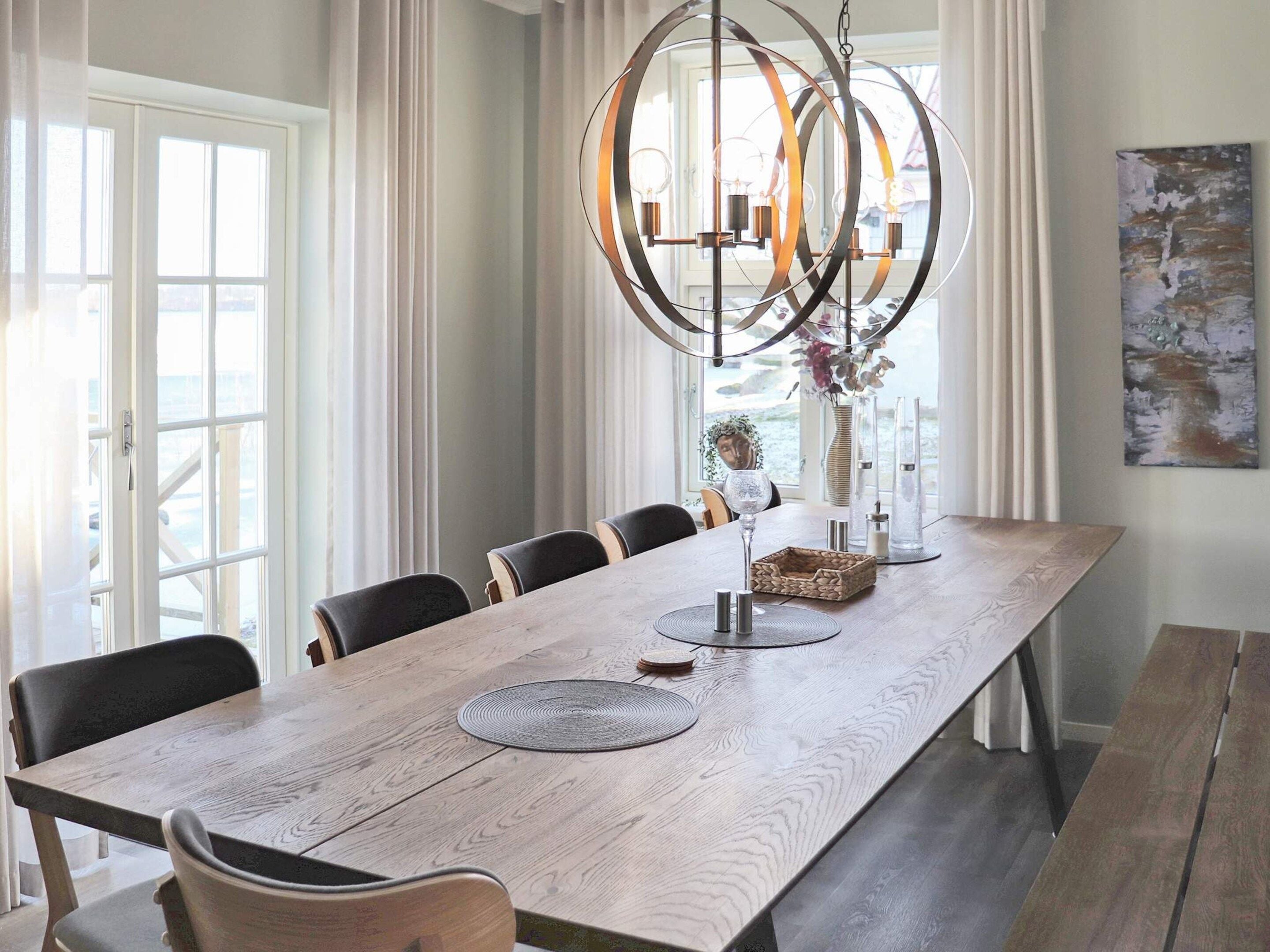 A dining room with a long wooden table, multiple chairs, a hanging light fixture, and a view through the window.