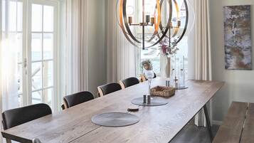 A dining room with a long wooden table, multiple chairs, a hanging light fixture, and a view through the window.