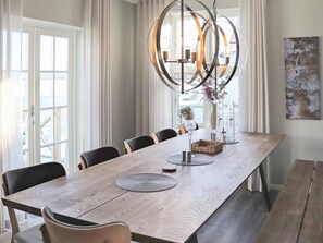 Une salle à manger avec une longue table en bois, plusieurs chaises, un luminaire suspendu et une vue par la fenêtre.