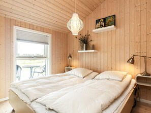 A bedroom with a wooden ceiling, a bed with white bedding, a hanging light fixture, a small table with a lamp, and a window with blinds. - 8 Person Holiday Home in Lokken (Lokken)