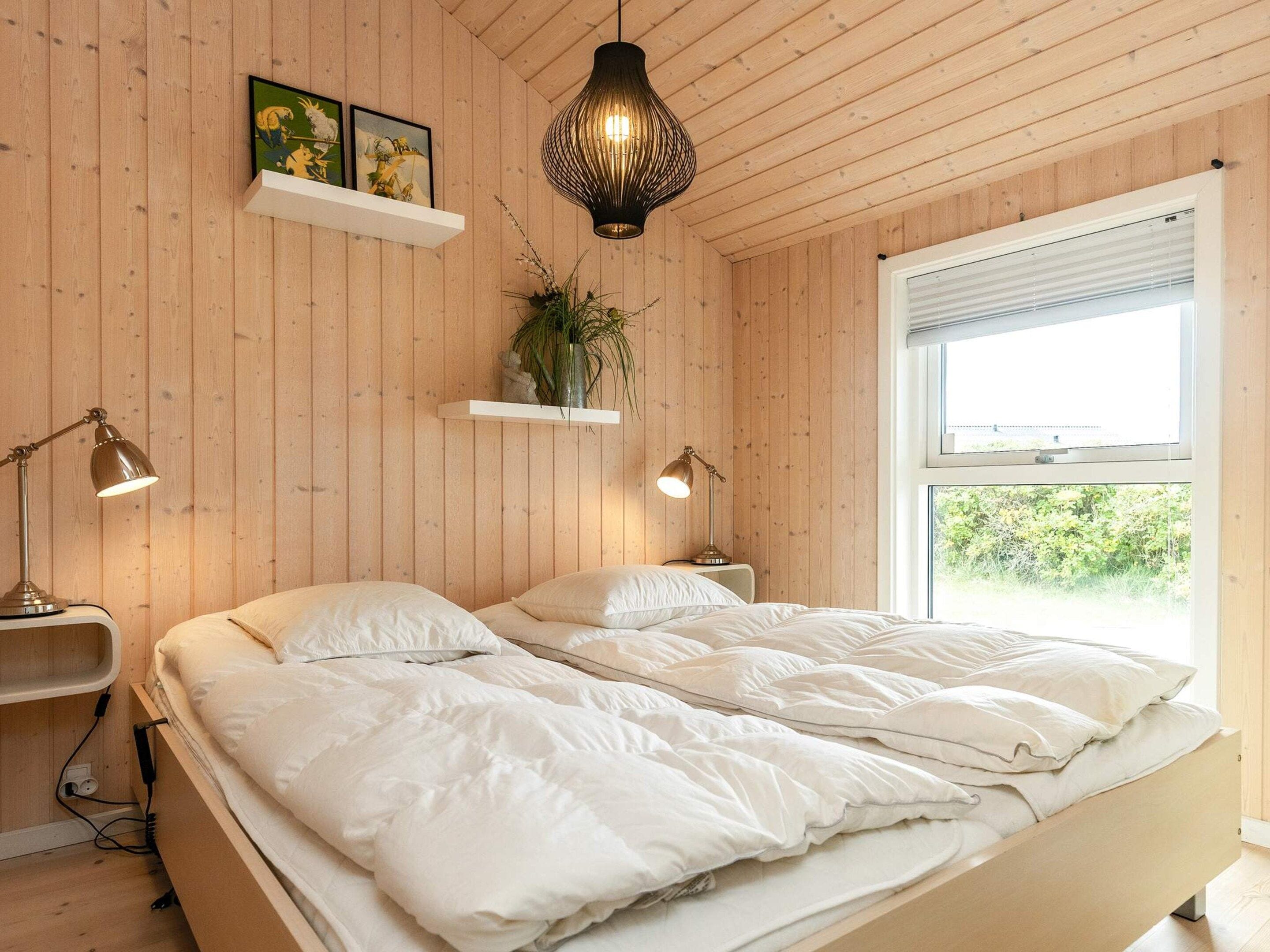 Un dormitorio con piso de madera, una cama con ropa blanca, dos lámparas de noche, una lámpara colgante y una ventana con persianas.