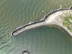 An aerial view of a coastal area with a curved stone breakwater, a sandy beach, and a small structure near the water's edge.
