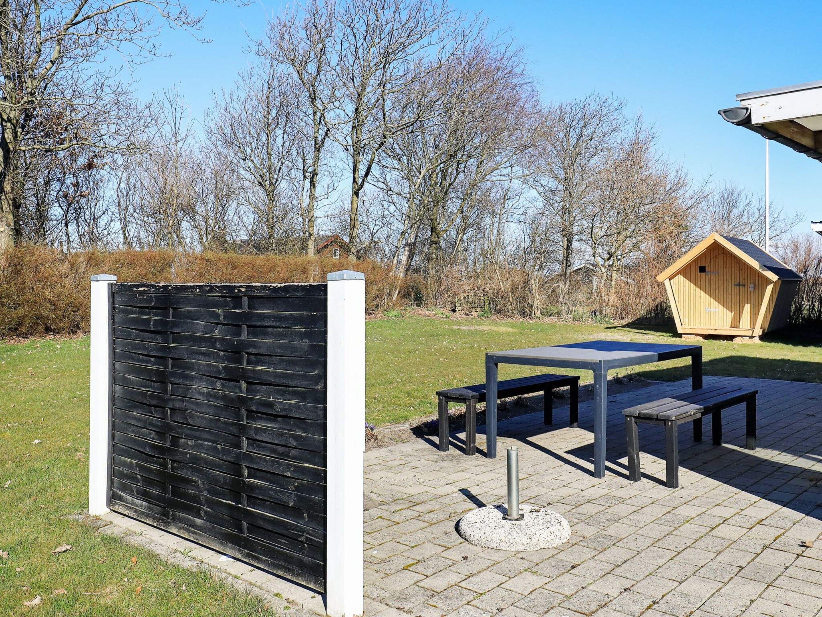 An outdoor seating area with a table and benches, a wooden structure, and a black fence.