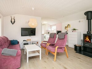 Un salon moderne avec un canapé, des fauteuils, un téléviseur, une cheminée et une cuisine en arrière-plan.