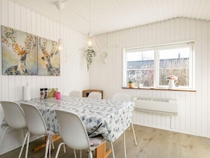 A dining area with a table set for four, white chairs, a patterned tablecloth, and a window with a view of a building and plants.