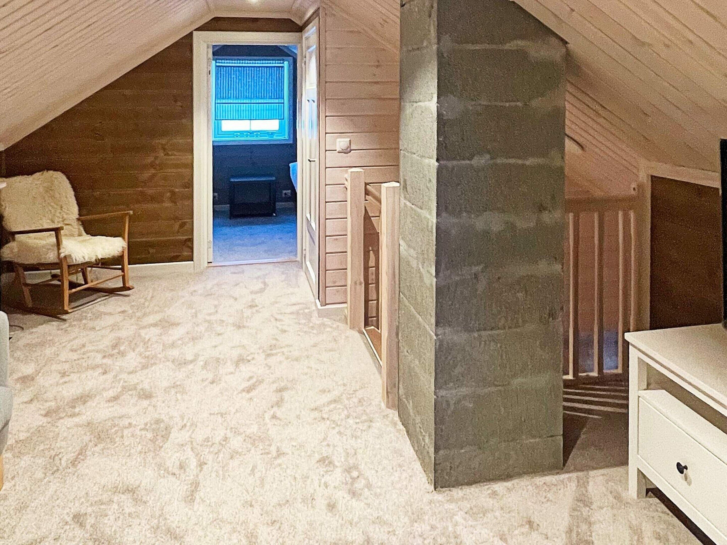 A cozy attic bedroom with a stone fireplace, a wooden chair, and a view through a window.