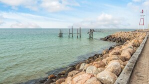 A coastal scene with a wooden pier extending into calm waters, a rocky breakwater, and a lighthouse in the distance. - 6 Person Holiday Home in Laeso-by Traum (Læsø)