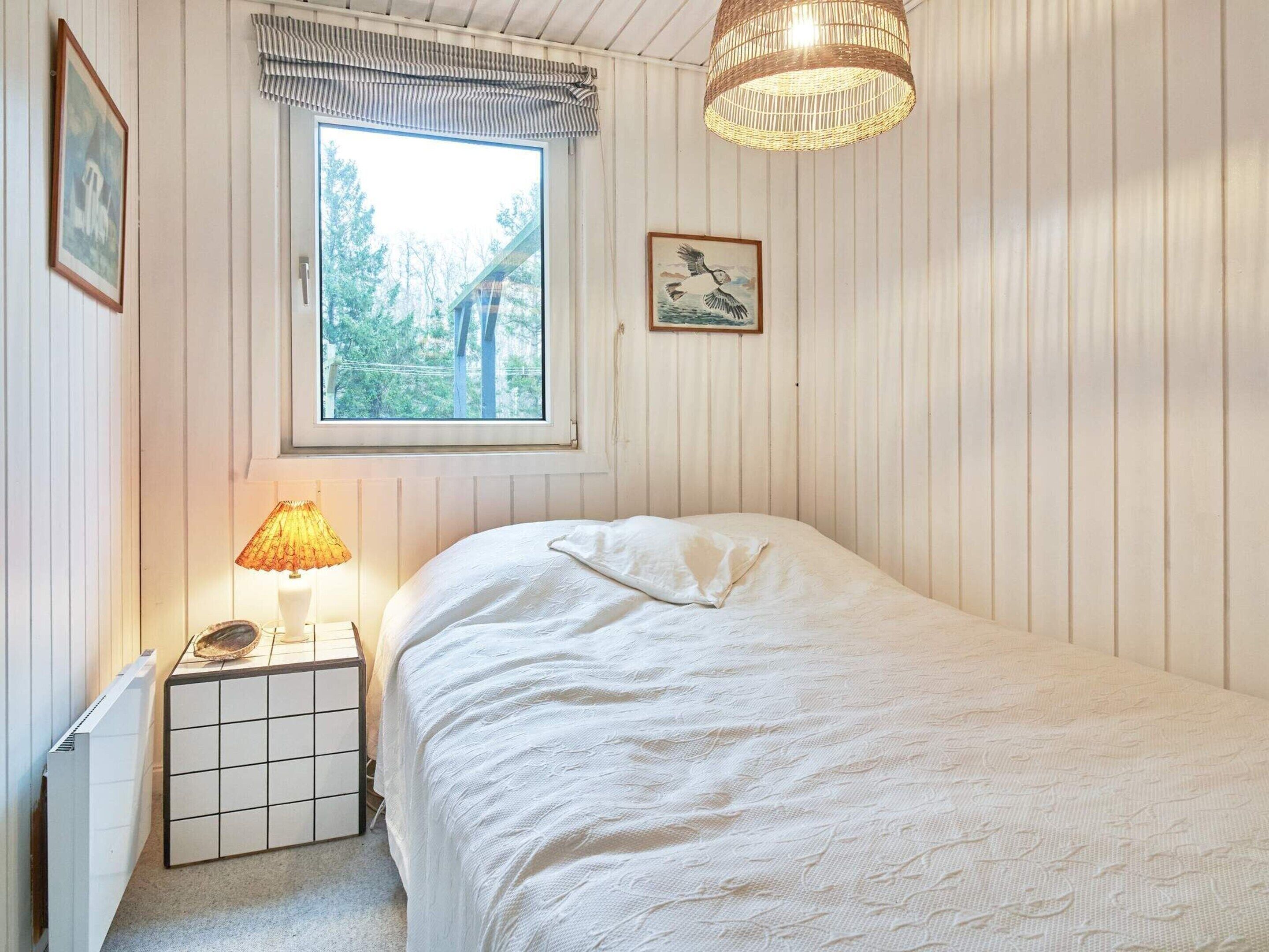 Ein Schlafzimmer mit großem Fenster, Nachttischlampe, einem Bett mit weißer Bettdecke und einem kleinen Nachttisch.