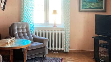 A living room with a sofa, a table, a radiator, a TV stand, and a window with curtains.