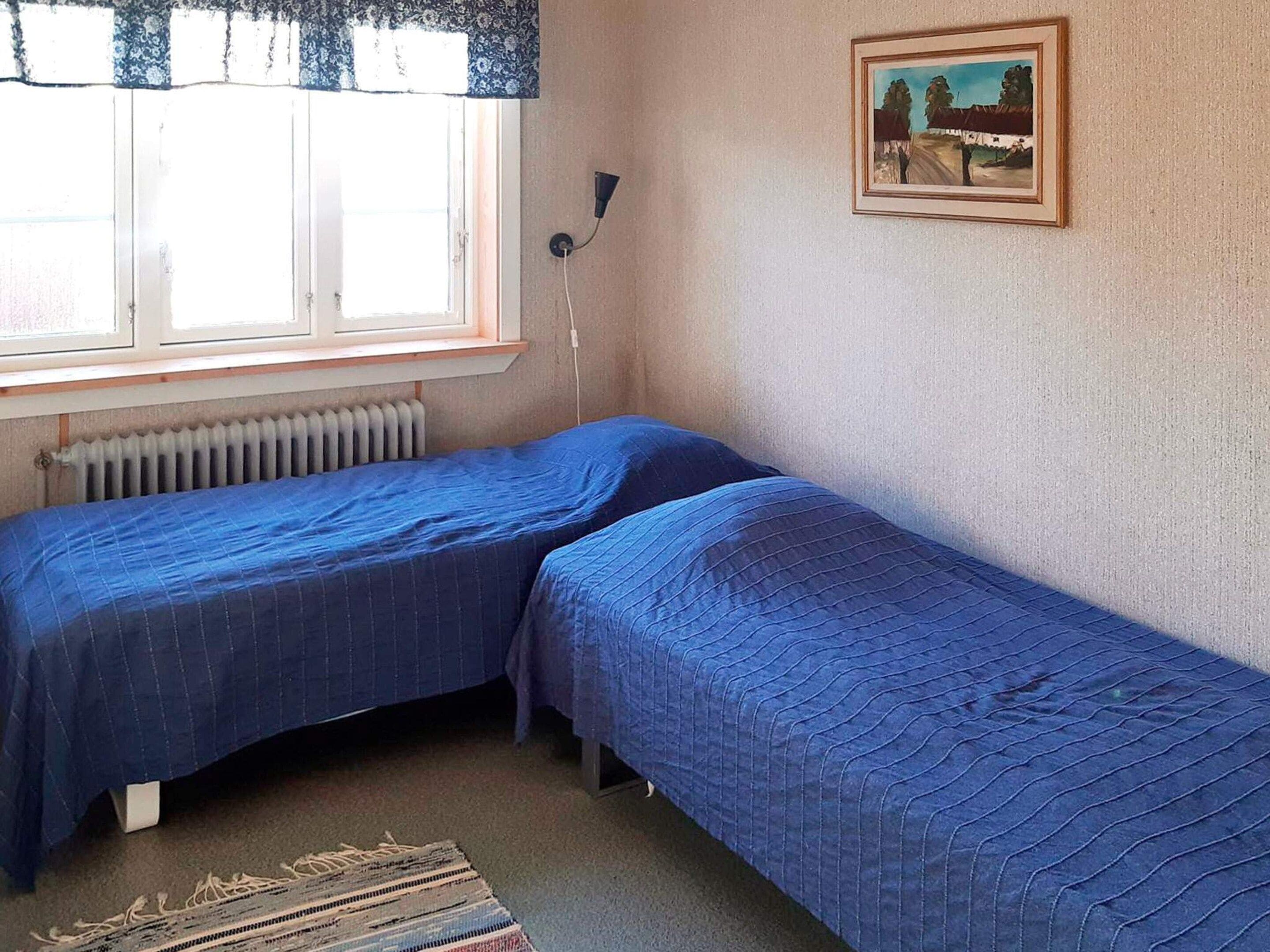 A room with two single beds, a radiator, a window with curtains, a wall-mounted lamp, and a framed picture.