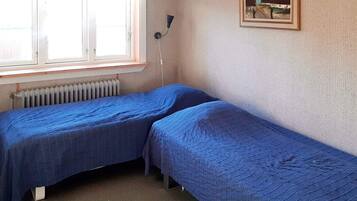 A room with two single beds, a radiator, a window with curtains, a wall-mounted lamp, and a framed picture.
