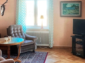 A living room with a sofa, a table, a radiator, a TV stand, and a window with curtains.