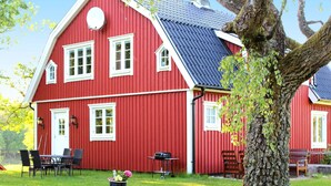 Ein rotes Holzhaus mit geneigtem Dach, ein großer Baum davor und eine Rasenfläche.