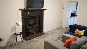 A living room with a fireplace, a flat-screen TV mounted above it, a sofa, and a small side table with a lamp. - Chapel Lodge by Tŷ SA (Newport)