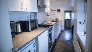A narrow kitchen with white cabinets, a microwave, a kettle, and a stove. - Chapel Lodge by Tŷ SA (Newport)