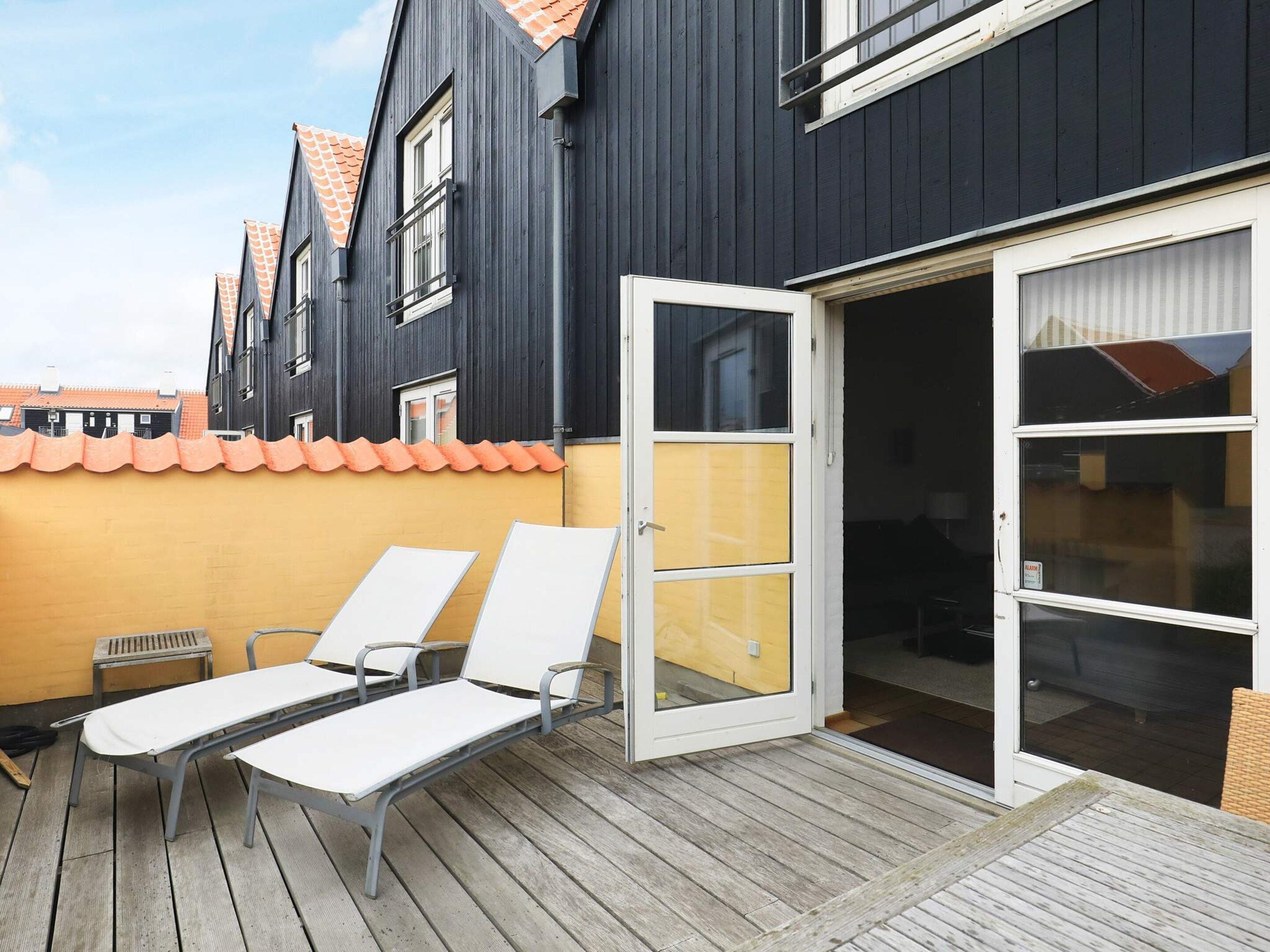 Eine Terrasse mit zwei weißen Liegestühlen und einem kleinen Tisch, eine Glastür zu einem überdachten Patio und ein Gebäude mit dunklen Wänden und orangefarbenen Dachziegeln.