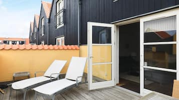 Une terrasse avec deux chaises longues blanches et une petite table, une porte-fenêtre coulissante donnant sur un patio abrité, et un bâtiment de couleur sombre avec un mur jaune et des tuiles de toit orange.