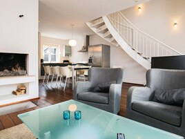 A modern living room with a glass coffee table, two grey armchairs, a fireplace, and a staircase leading to an upper level.