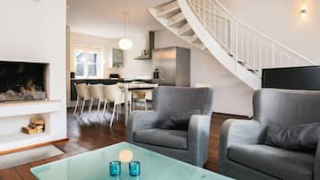 A modern living room with a glass coffee table, two grey armchairs, a fireplace, and a staircase.