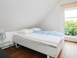 Una cama individual con colcha blanca y azul, una lámpara de noche y una ventana con vistas a follaje.
