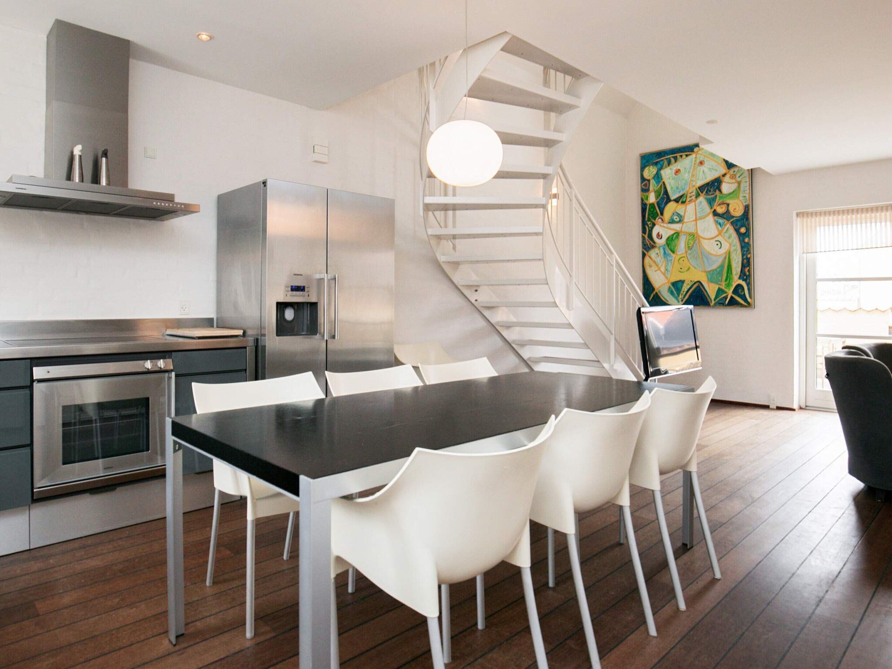 Cozinha moderna com eletrodomésticos de aço inoxidável, área de jantar com mesa preta e cadeiras brancas, e escada em espiral que leva a um andar superior.