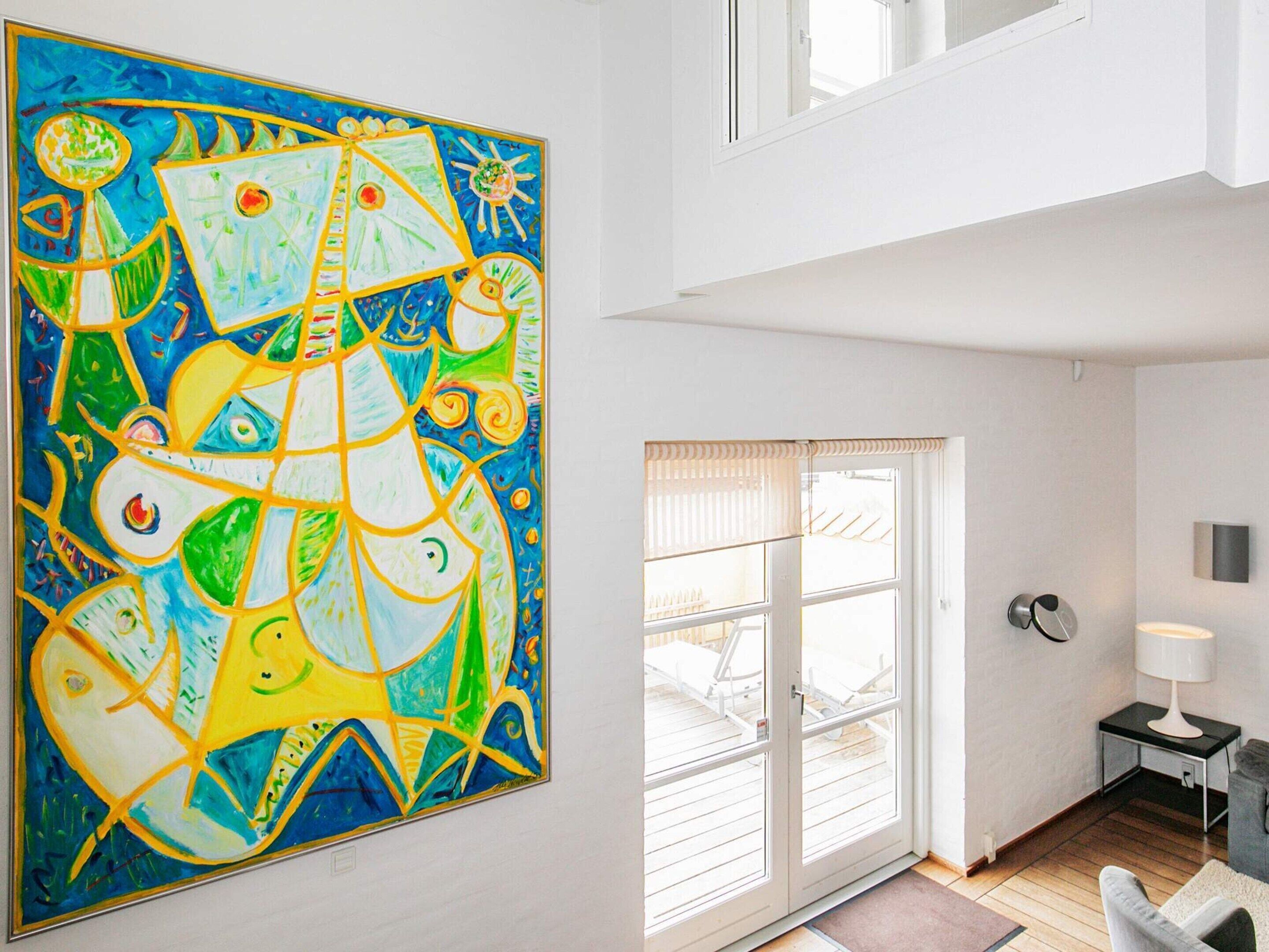 Quarto com uma grande pintura abstrata na parede, porta de vidro deslizante que dá para uma área externa e uma pequena mesa com abajur.