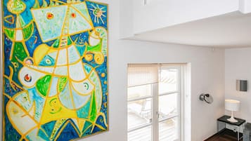 Un interior moderno con un cuadro abstracto grande en la pared, una puerta de vidrio corrediza que da a un espacio exterior, y una mesita de noche con una lámpara.