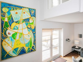 Un interior moderno con un cuadro abstracto grande en la pared, una puerta de vidrio corrediza que da a un espacio exterior, y una mesita de noche con una lámpara.