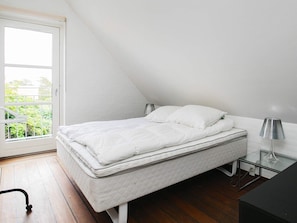 A single bed in a room with a sloped ceiling, a glass table with a lamp, and a view of greenery outside through a window. - 4 Star Holiday Home in Skagen (Skagen)