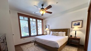 Une chambre à coucher comprenant un lit, une table de chevet, un ventilateur de plafond, une fenêtre avec des stores et un tableau encadré accroché au mur.