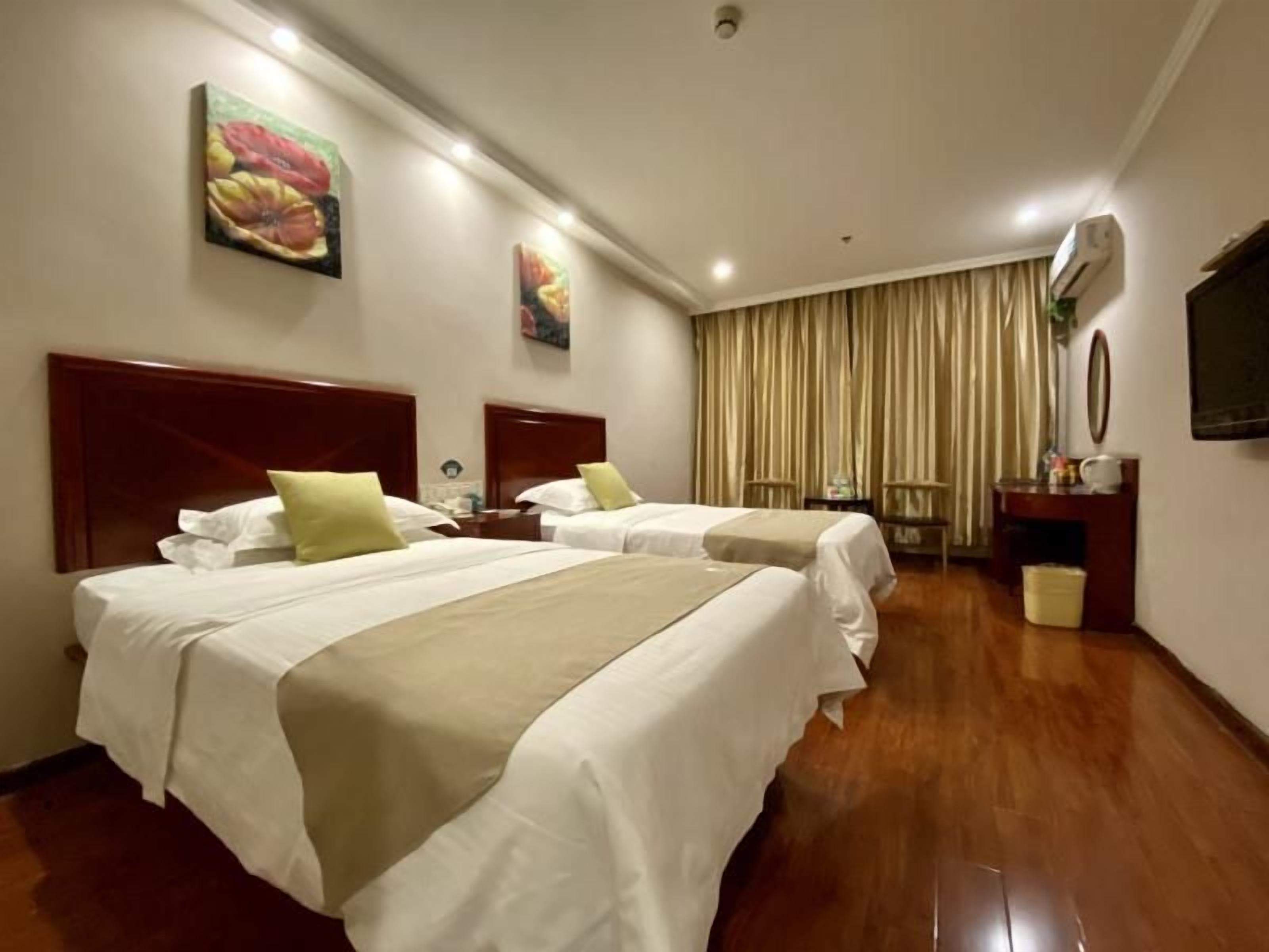 A hotel room with two beds, a wooden floor, a large window with curtains, and a flat-screen TV on the wall.