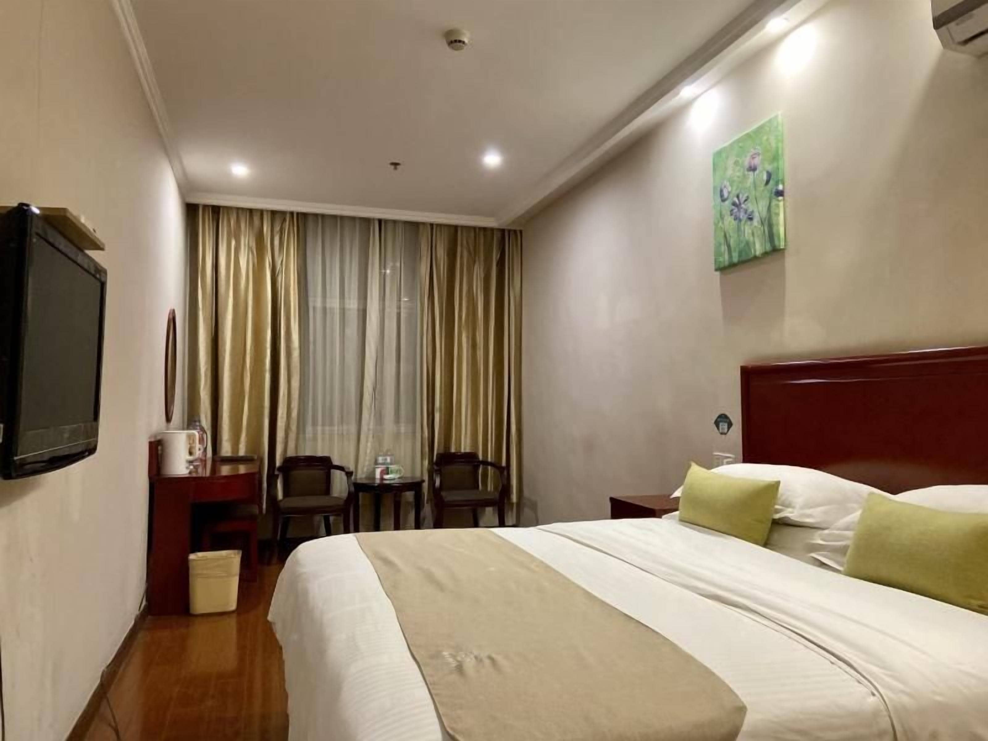 A hotel room with a bed, a television, a desk with a chair, a window with curtains, and a painting on the wall.