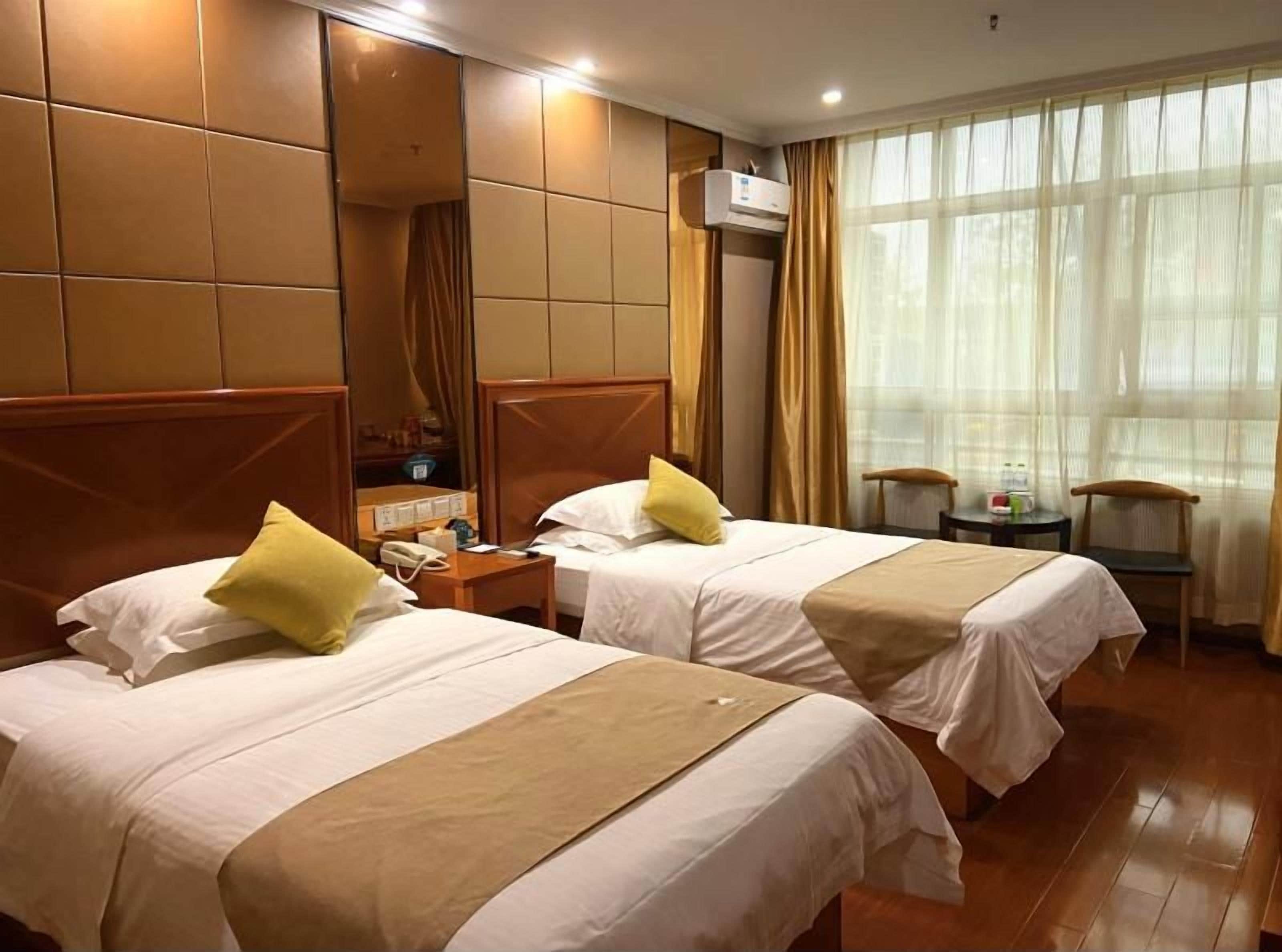 A hotel room with two beds, a wooden headboard, an air conditioning unit, a small table with a chair, and a window with sheer curtains.