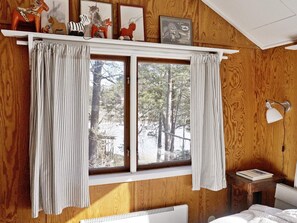A room with a bed, a bedside table with a lamp, a window with curtains, and wooden walls. - 6 Person Holiday Home in Djurhamn (Djurhamn)