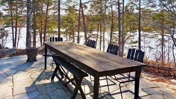 Una mesa de picnic de madera con bancos, ubicada en un patio de piedra con vistas a un bosque y una masa de agua.