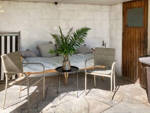 Een klein terras met een bed, twee rieten stoelen, een tafel met een bloemenvaas en een houten deur.