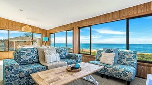 A spacious living area with large windows overlooking the ocean, a wooden ceiling, and a central coffee table.