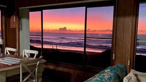 A room with a large window overlooking the ocean at sunset, a wooden dining table, and a patterned sofa.
