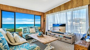 A living room with a large TV, wooden walls, a sofa, and a view of the ocean.