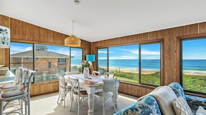 A spacious room with a large window overlooking a beach, a dining area with white furniture, and a view of a stone building.