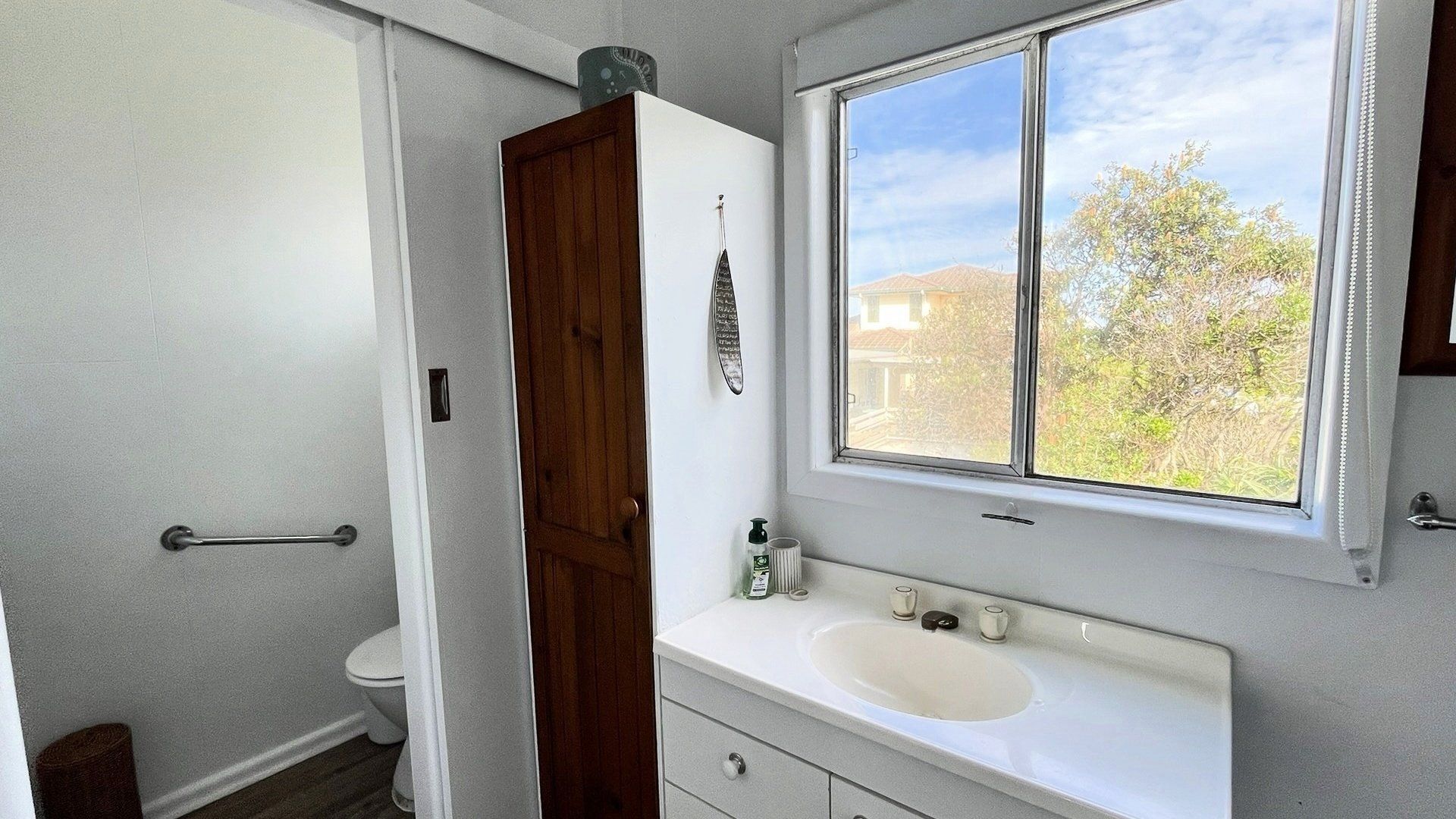 Een badkamer met een witte wastafel, een houten deur en een raam met uitzicht op huizen en bomen.