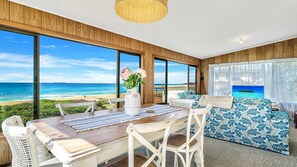 A room with a large window overlooking a beach, a dining table with chairs, and a sofa with a view.