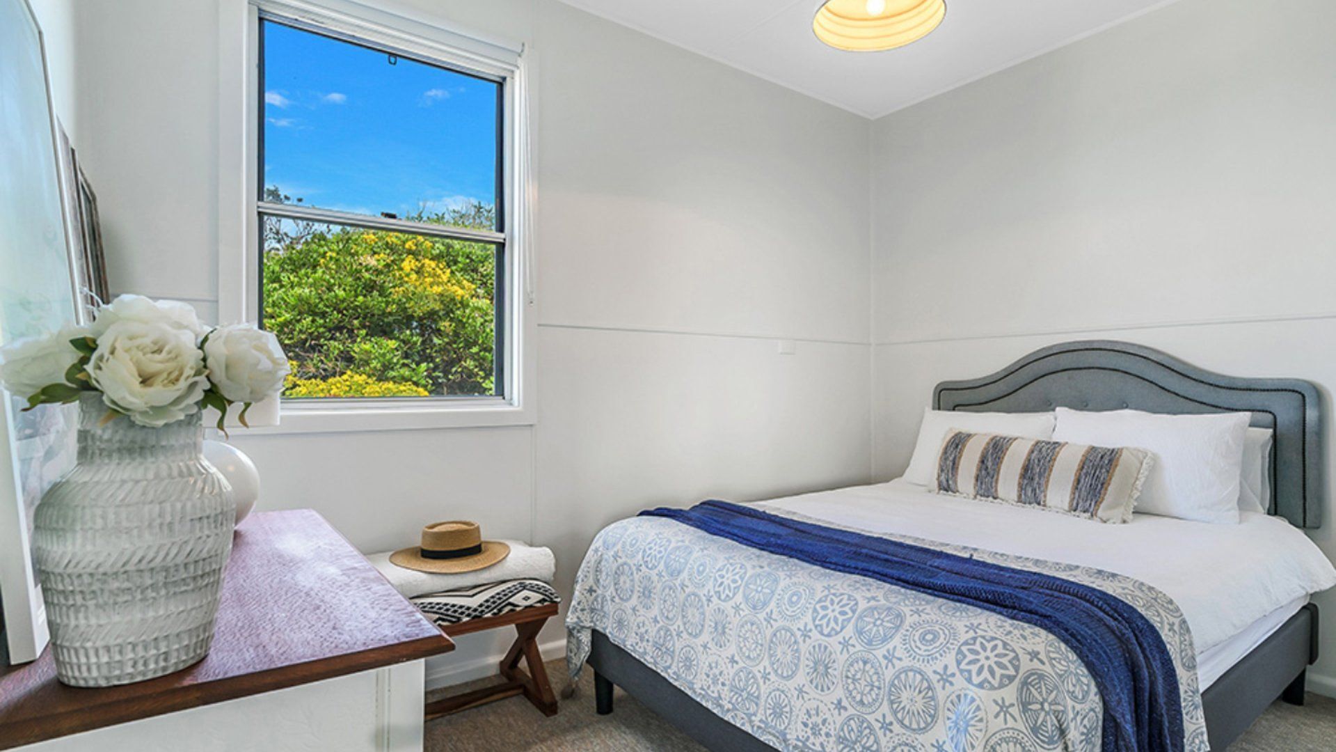 A bedroom with a bed, a nightstand, a vase with flowers, and a window with a view of greenery.