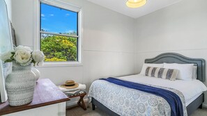 A bedroom with a bed, a nightstand, a vase with flowers, and a window with a view of greenery.