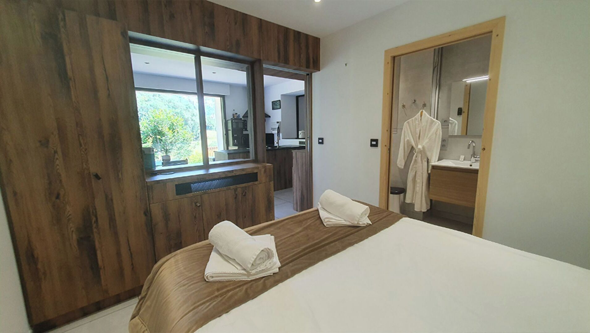 A modern bedroom with a large bed, wooden wardrobe, a mirror, and a view of the kitchen.