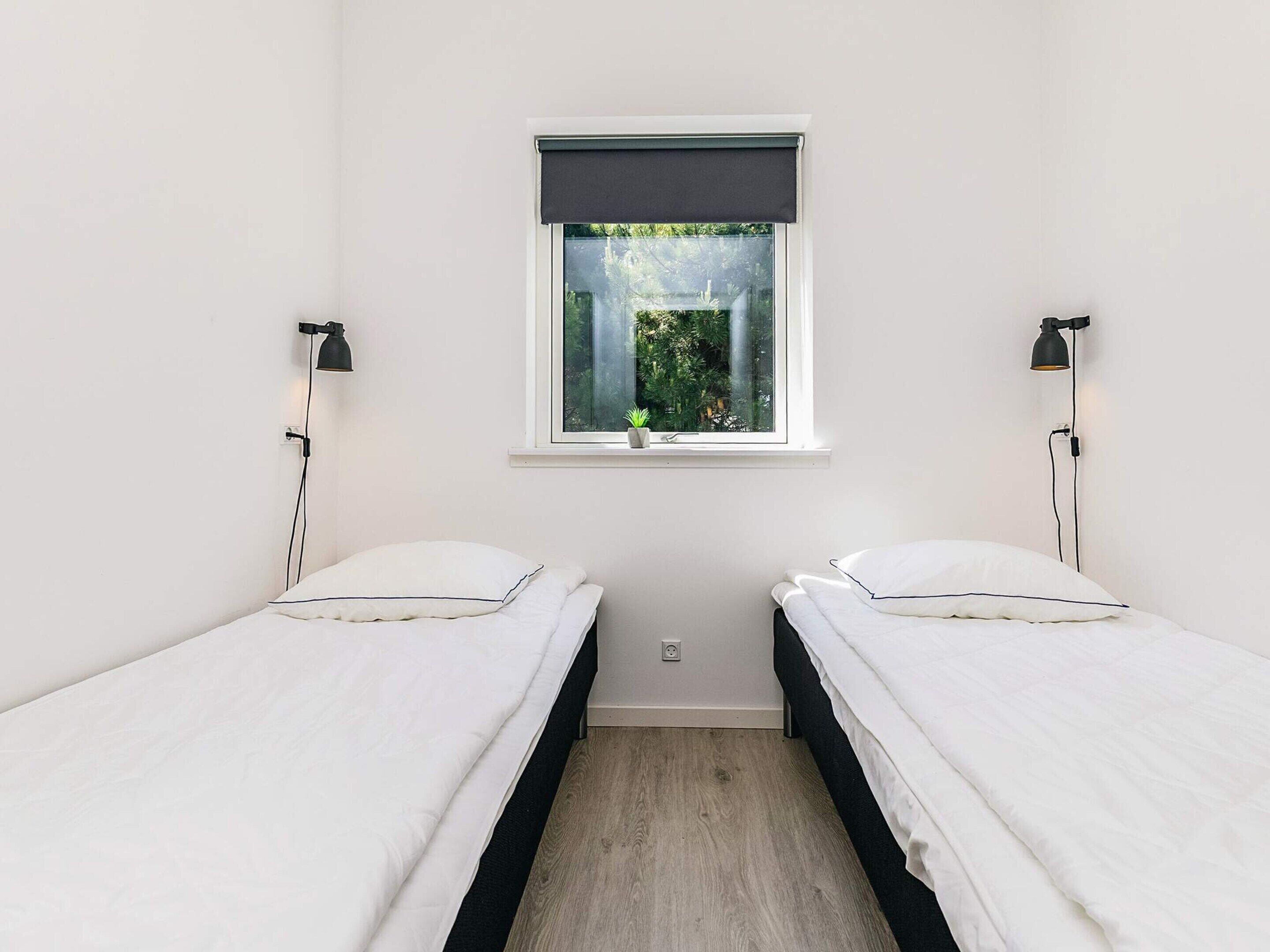 A small, clean room with two single beds, a window with a view of greenery, and two wall-mounted reading lamps.